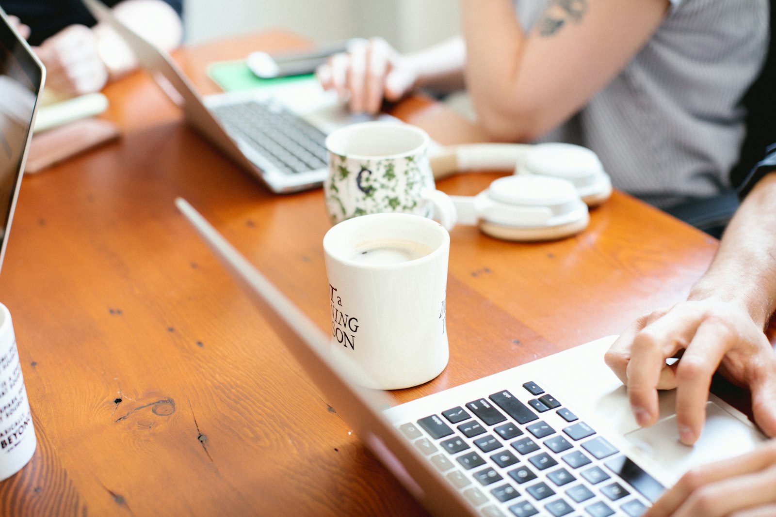 Two people working side by side on laptops at a wooden table with coffee mugs